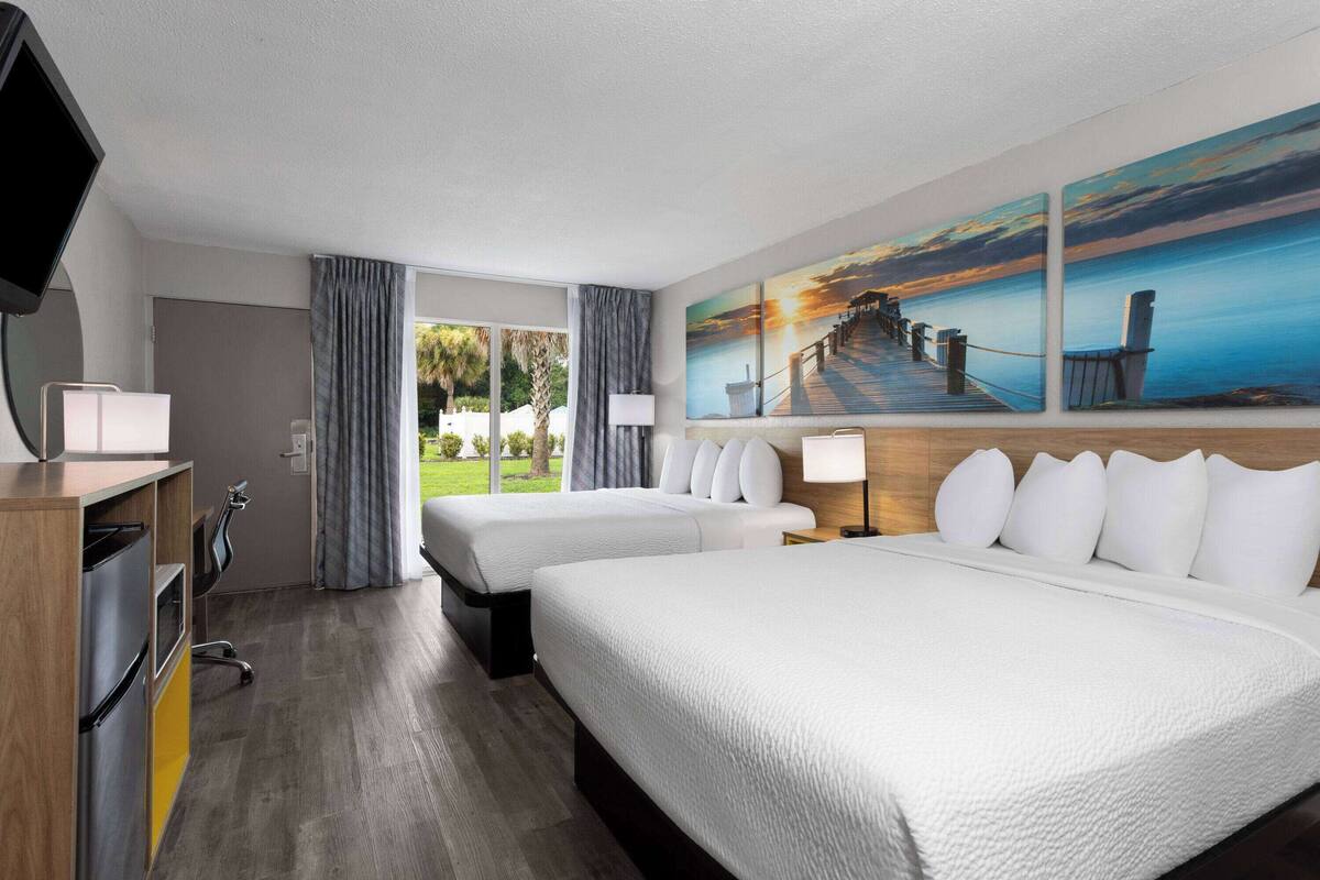 A hotel room with two beds, a TV, a desk, and a painting of a sunset over a pier.