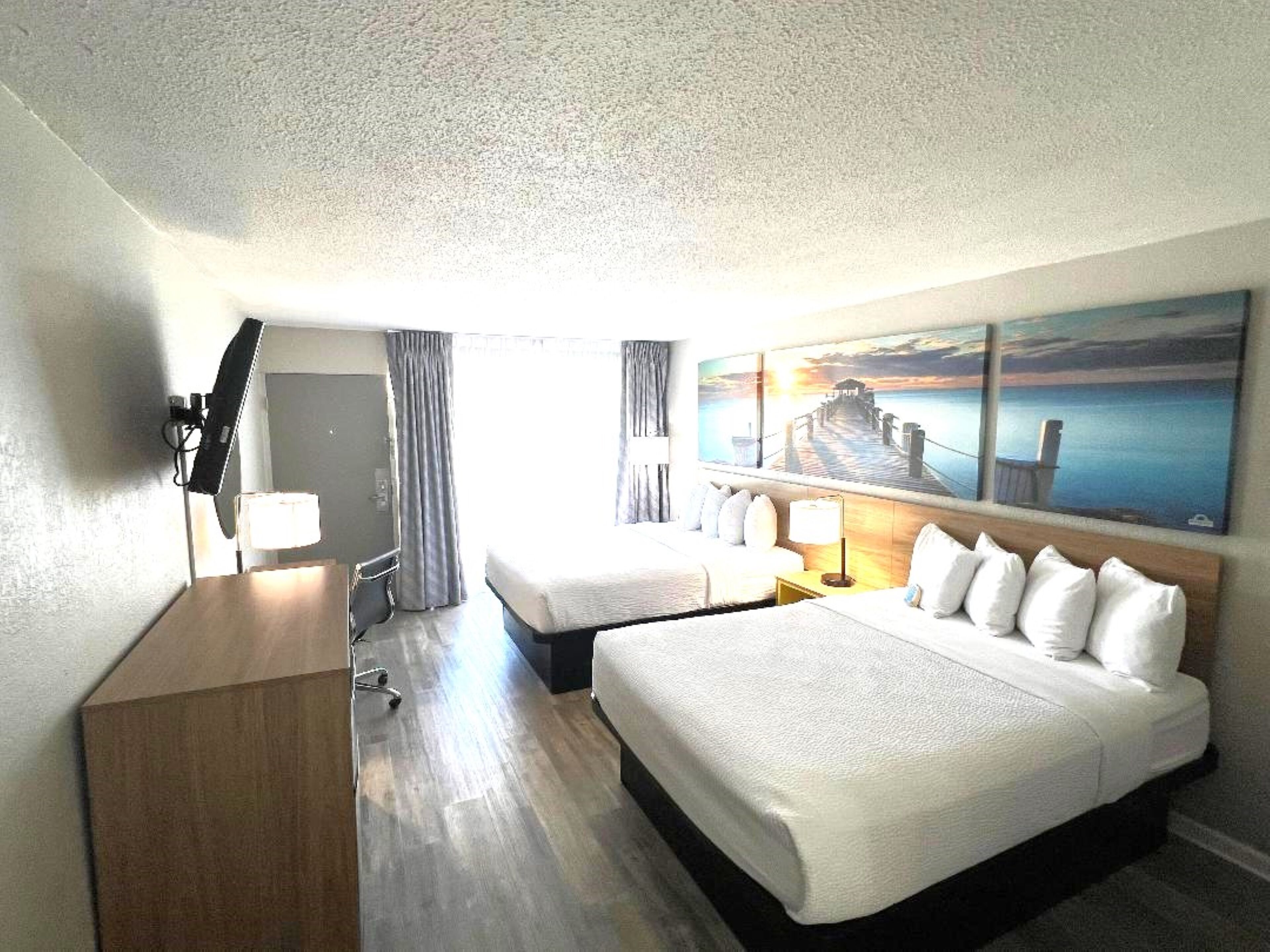 A hotel room with two beds, a desk, a chair, a TV, and a painting of a pier.