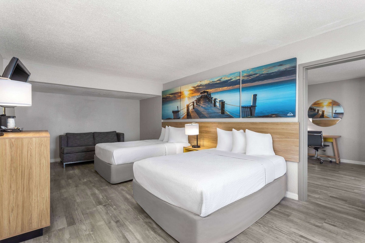 A hotel room with a large bed, two smaller beds, a desk, a chair, a lamp, and a painting of a sunset over a pier.