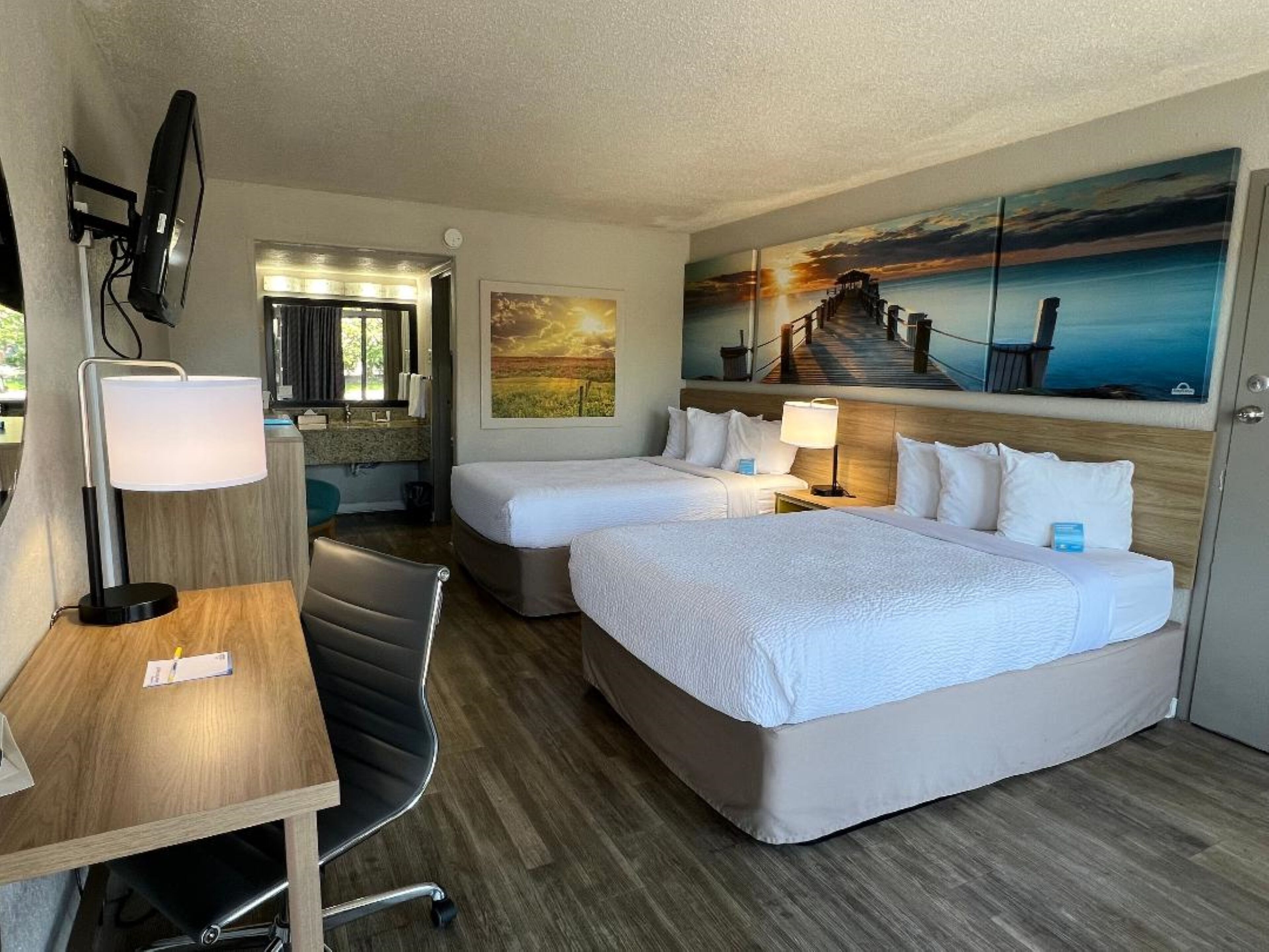Habitación de hotel con dos camas, un escritorio con lámpara, una silla, un televisor y un cuadro de un atardecer sobre un muelle.