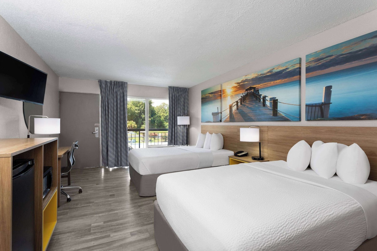 A hotel room with two beds, a desk, a TV, and a balcony with curtains.