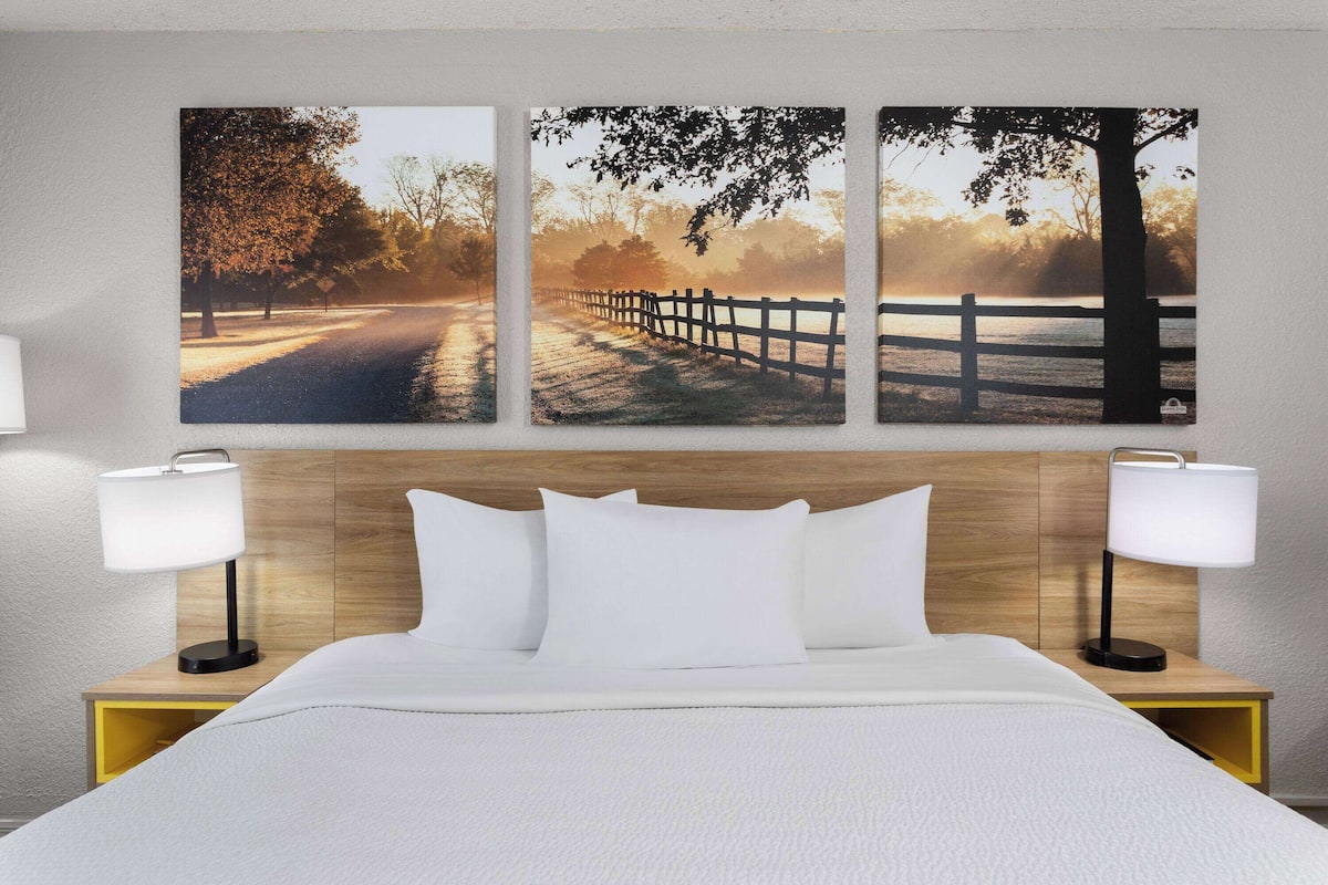 A hotel room with a wooden headboard, white bedding, and three landscape paintings on the wall.