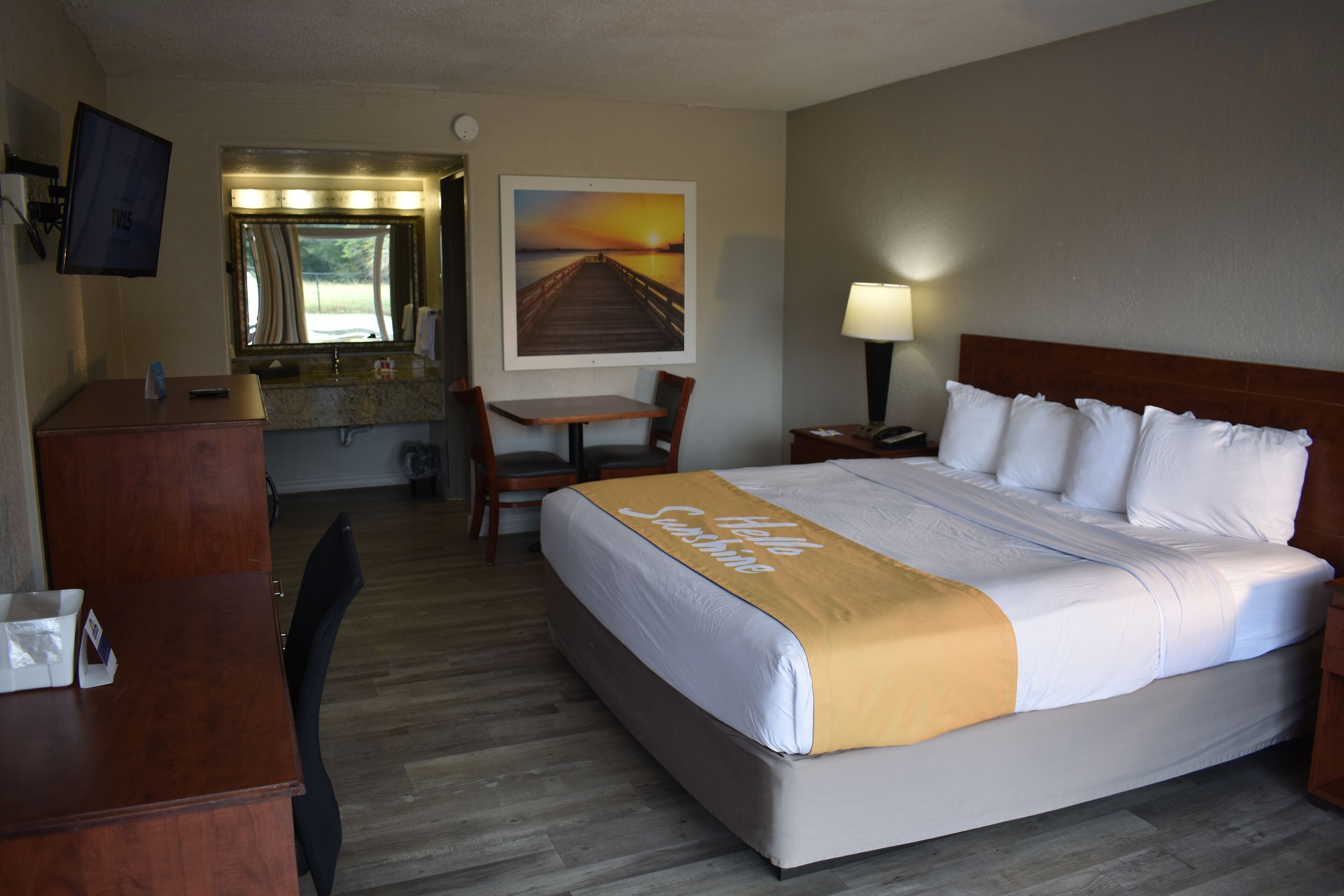 A hotel room with a bed, a desk, a chair, a TV, a bathroom with a mirror, and a painting of a sunset.
