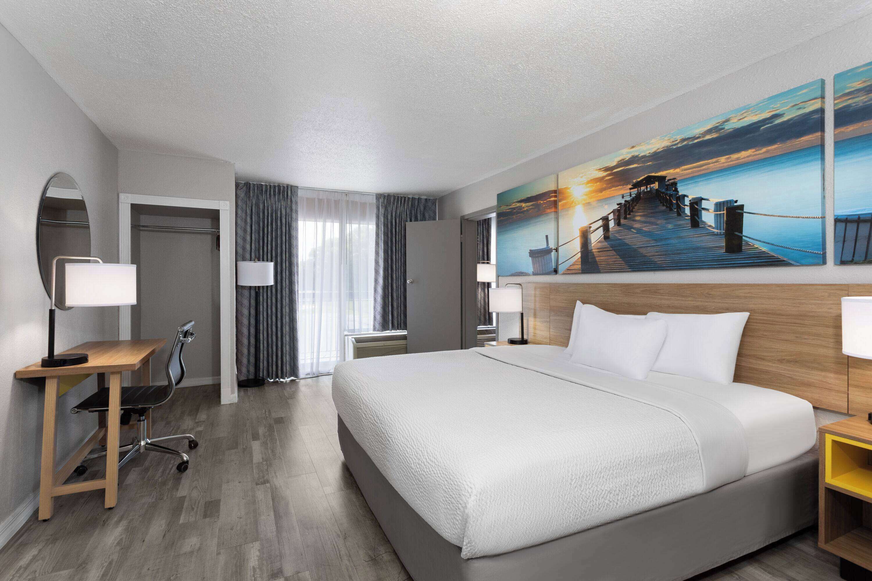 A hotel room with a large bed, a desk, a chair, a desk lamp, a nightstand, a painting of a pier, and a window with curtains.