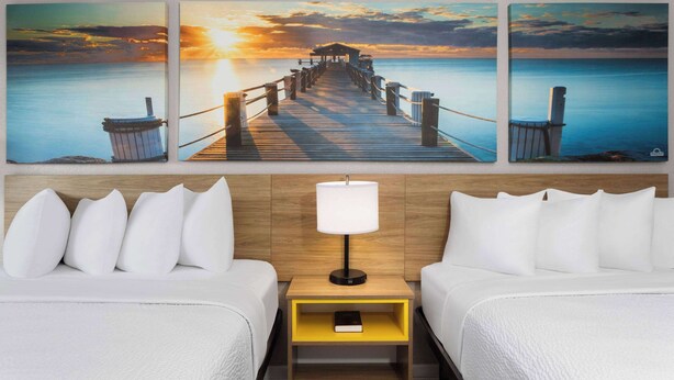 A hotel room with two beds, a nightstand with a lamp, and a wall-mounted painting of a pier and sunset. - Days Inn by Wyndham Wildwood I-75 (Wildwood)