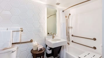 A modern bathroom with a white sink, toilet, and bathtub. There are towel racks and a small table with a towel and a tissue box.