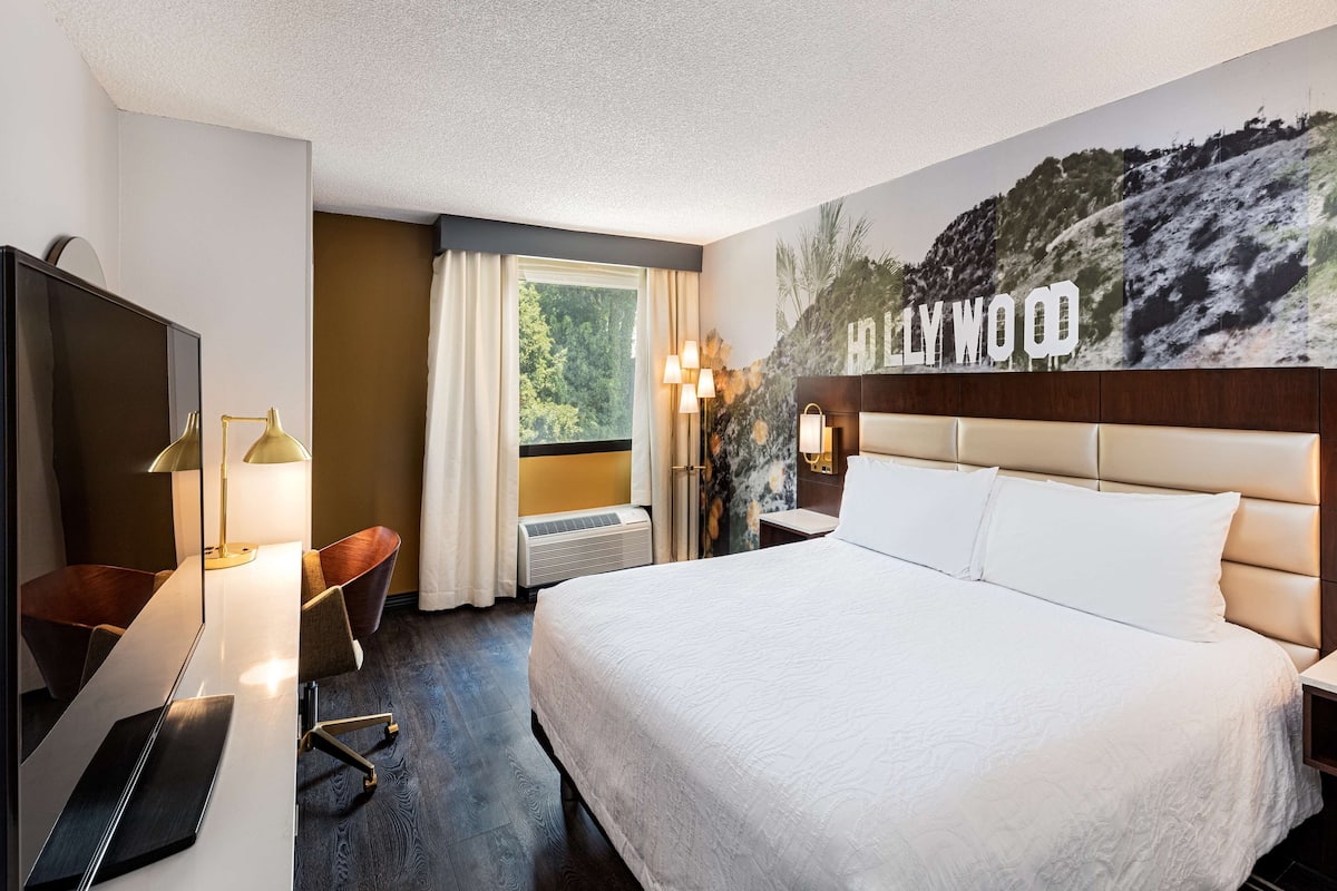 A hotel room with a large bed, a TV, a desk with a lamp, and a view of a mountain.