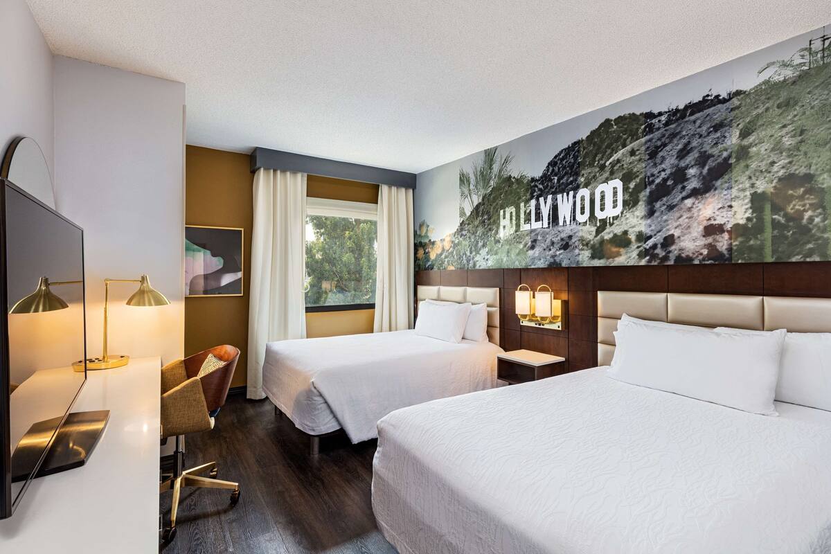 A hotel room with two beds, a desk with a lamp, a chair, a large window, and a wall mural.