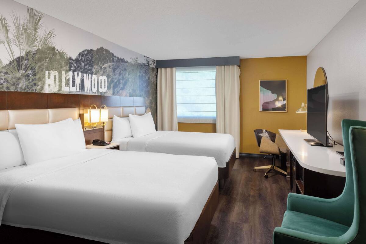 A hotel room with two beds, a desk, a chair, a television, and a mural of Hollywood.