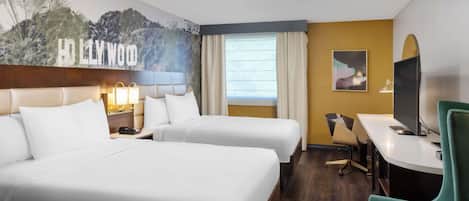 A hotel room with two beds, a desk, a chair, a television, and a mural of Hollywood.