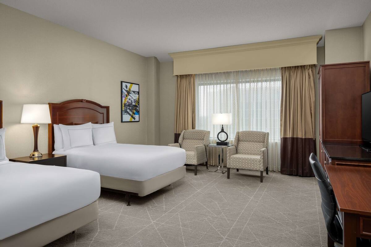 Une chambre d’hôtel avec deux lits, un bureau, une chaise, une télévision et une fenêtre avec des rideaux.
