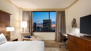 A hotel room with a large bed, a desk, a chair, a TV, and a view of a cityscape at dusk. - Hilton New York Times Square (New York)