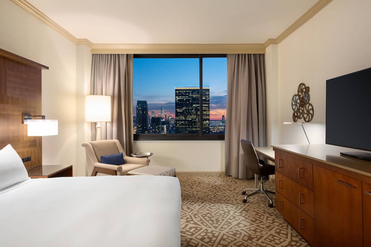 Une chambre d’hôtel avec un grand lit, un bureau, une chaise, une télévision et une vue sur la ville au crépuscule.