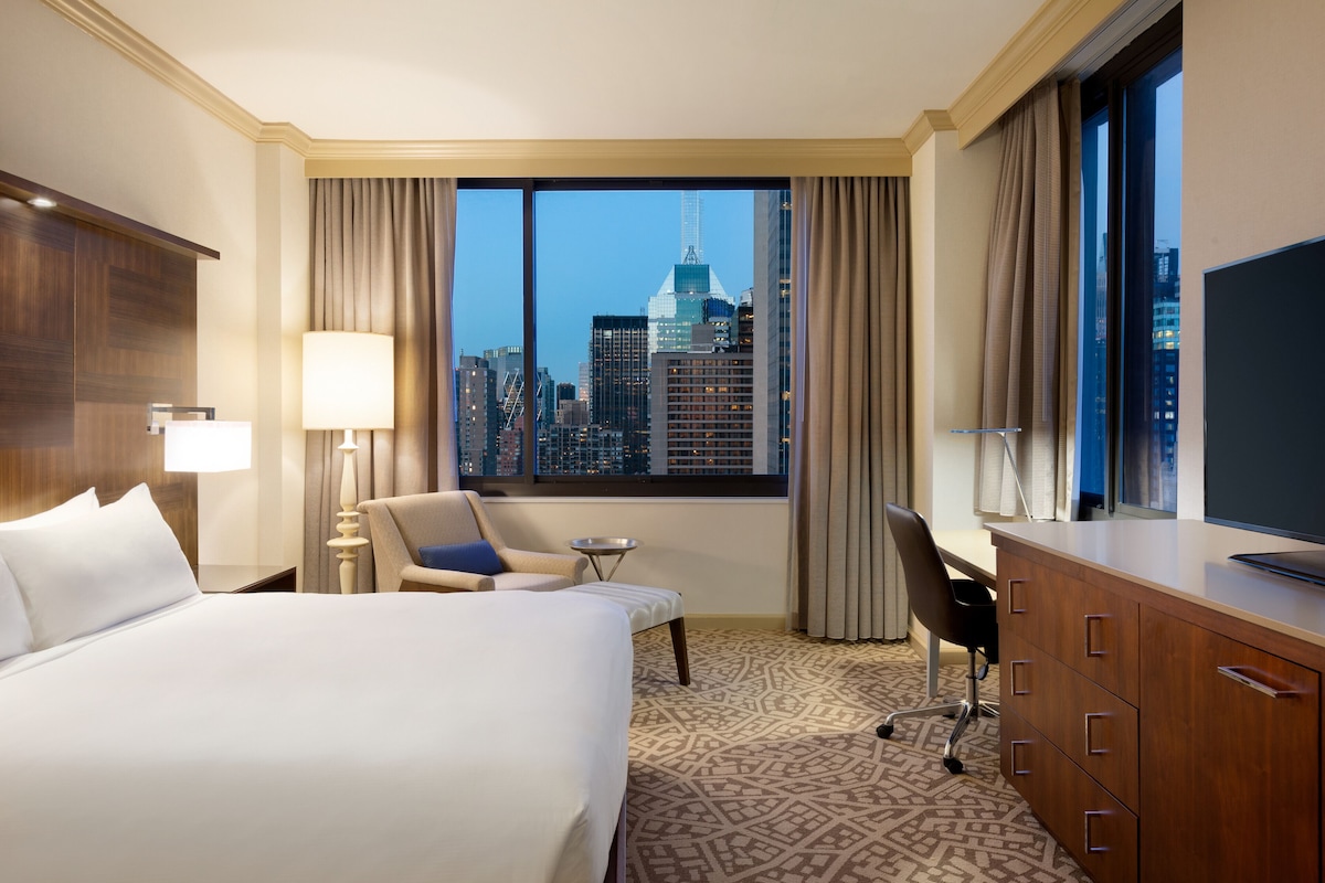 Une chambre d’hôtel avec un grand lit, un bureau, une chaise, une télévision et une vue sur le panorama de la ville.