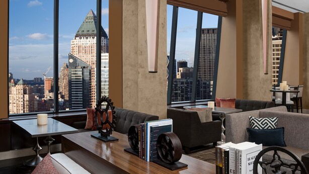 A modern hotel room with a large window offering a city view, a cozy seating area, and a small table with books and decorative items. - Hilton New York Times Square (New York)