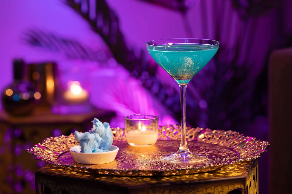 Un verre à cocktail contenant une boisson bleue, un petit bol de glaçons et une bougie posée sur un plateau décoratif.