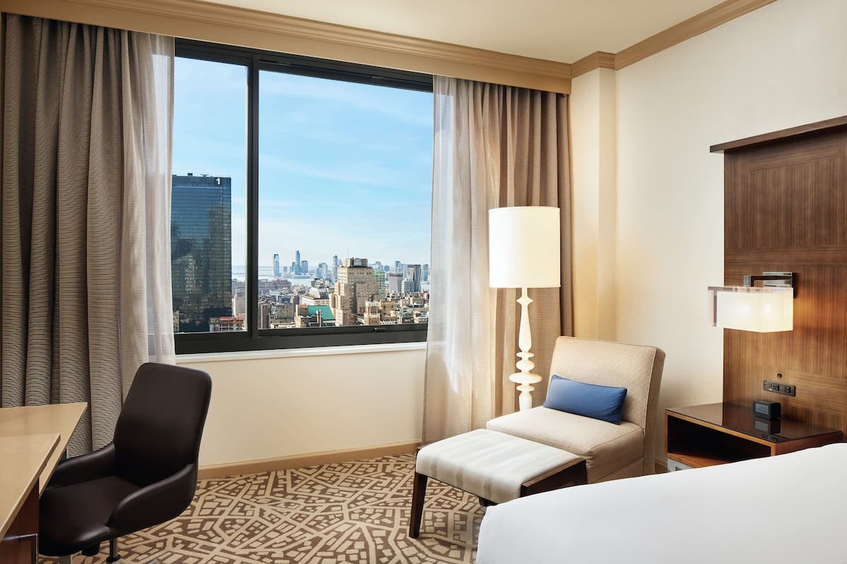 Une chambre d’hôtel équipée d’un lit, d’un bureau, d’une chaise, d’une lampe et offrant une vue sur le paysage urbain grâce à de grandes fenêtres.