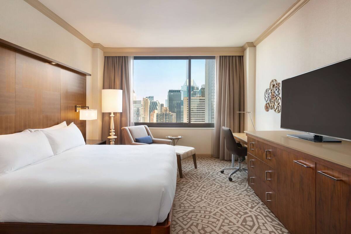 Une chambre d’hôtel dotée d’un grand lit, d’un bureau avec un téléviseur à écran plat, d’une chaise, d’une lampe et offrant une vue sur le panorama de la ville par une grande fenêtre.