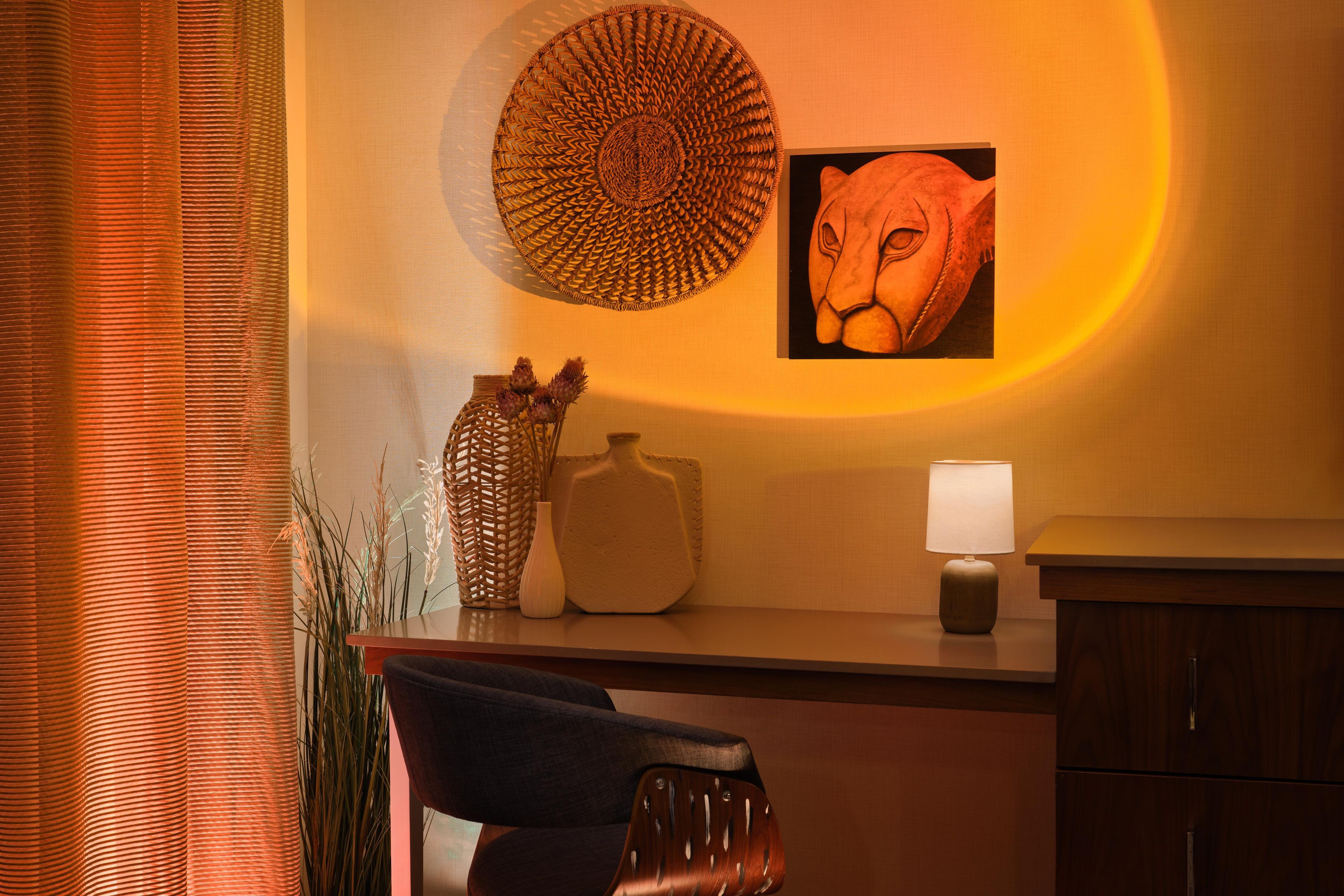 A room with a desk, a chair, a lamp, a woven basket, and a framed picture on the wall.
