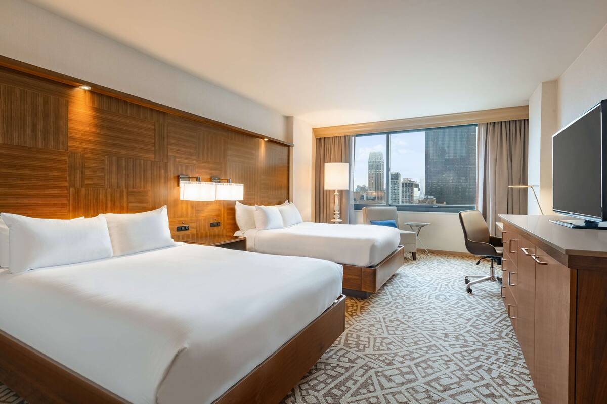 Une chambre d’hôtel avec deux lits, un bureau, une télévision et une grande fenêtre donnant sur la ville.