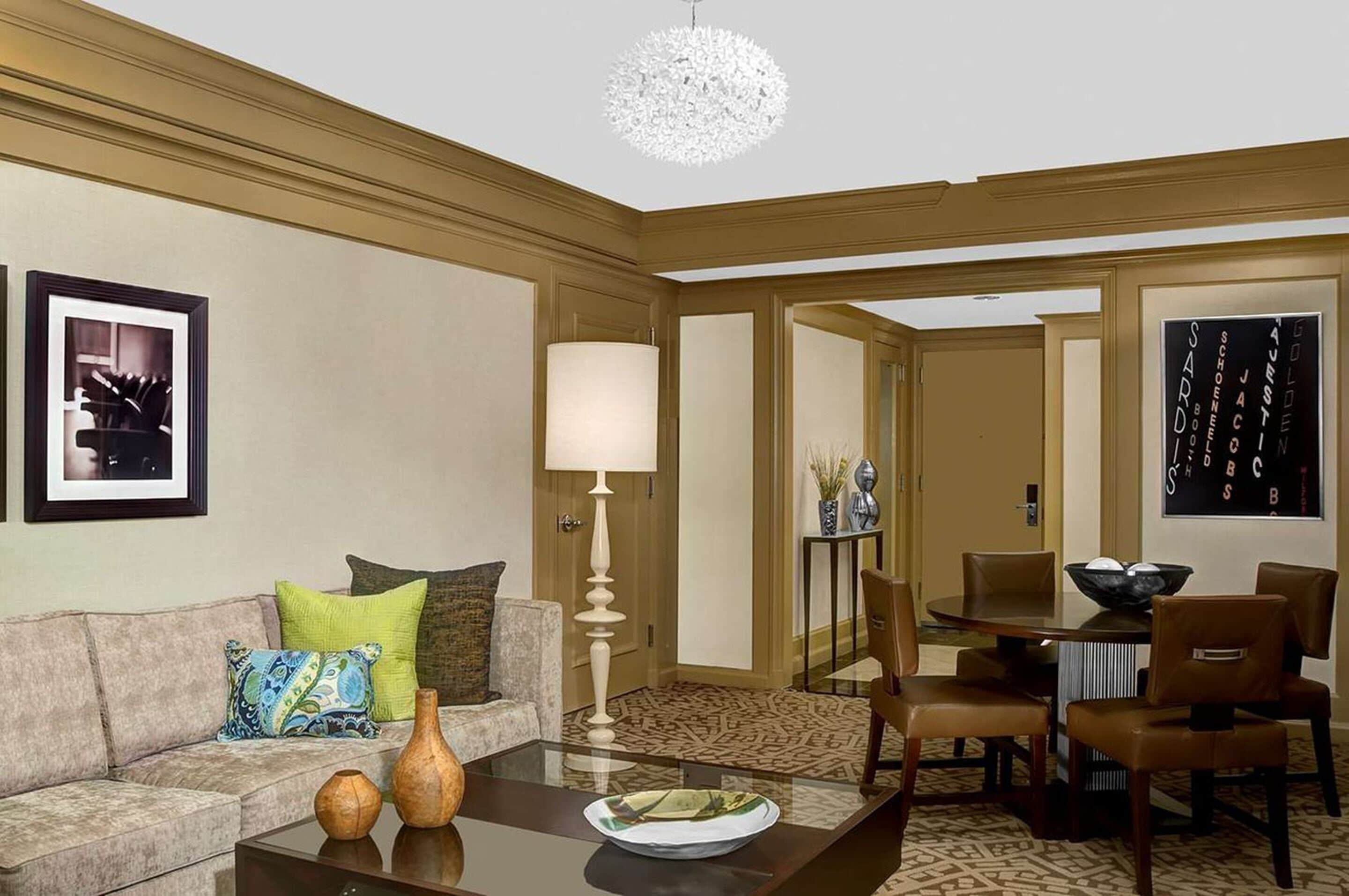 A hotel room with a sofa, a coffee table, a dining table with chairs, a chandelier, and a framed picture on the wall.