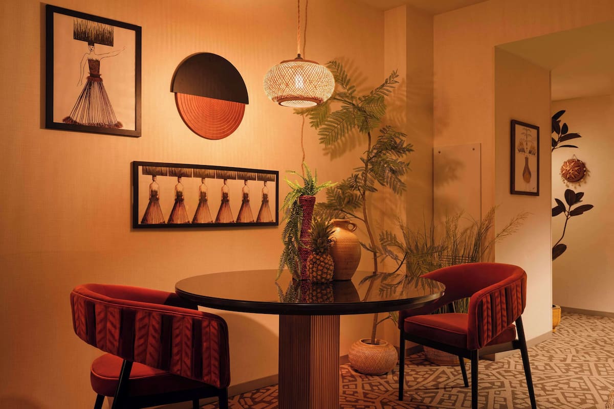 Un coin repas comprenant une table ronde, deux chaises à revêtement rouge, un luminaire suspendu en fibres tressées et des œuvres d’art accrochées au mur.