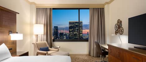 A hotel room with a large bed, a desk, a chair, a TV, and a view of a cityscape at dusk.