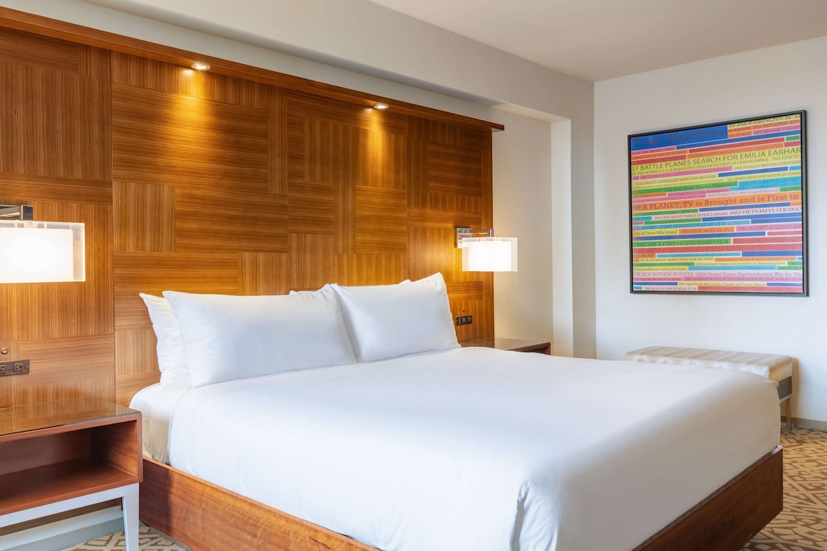 Une chambre d’hôtel avec un grand lit, une tête de lit en bois, deux lampes de chevet et une affiche colorée contenant des informations utiles accrochée au mur.