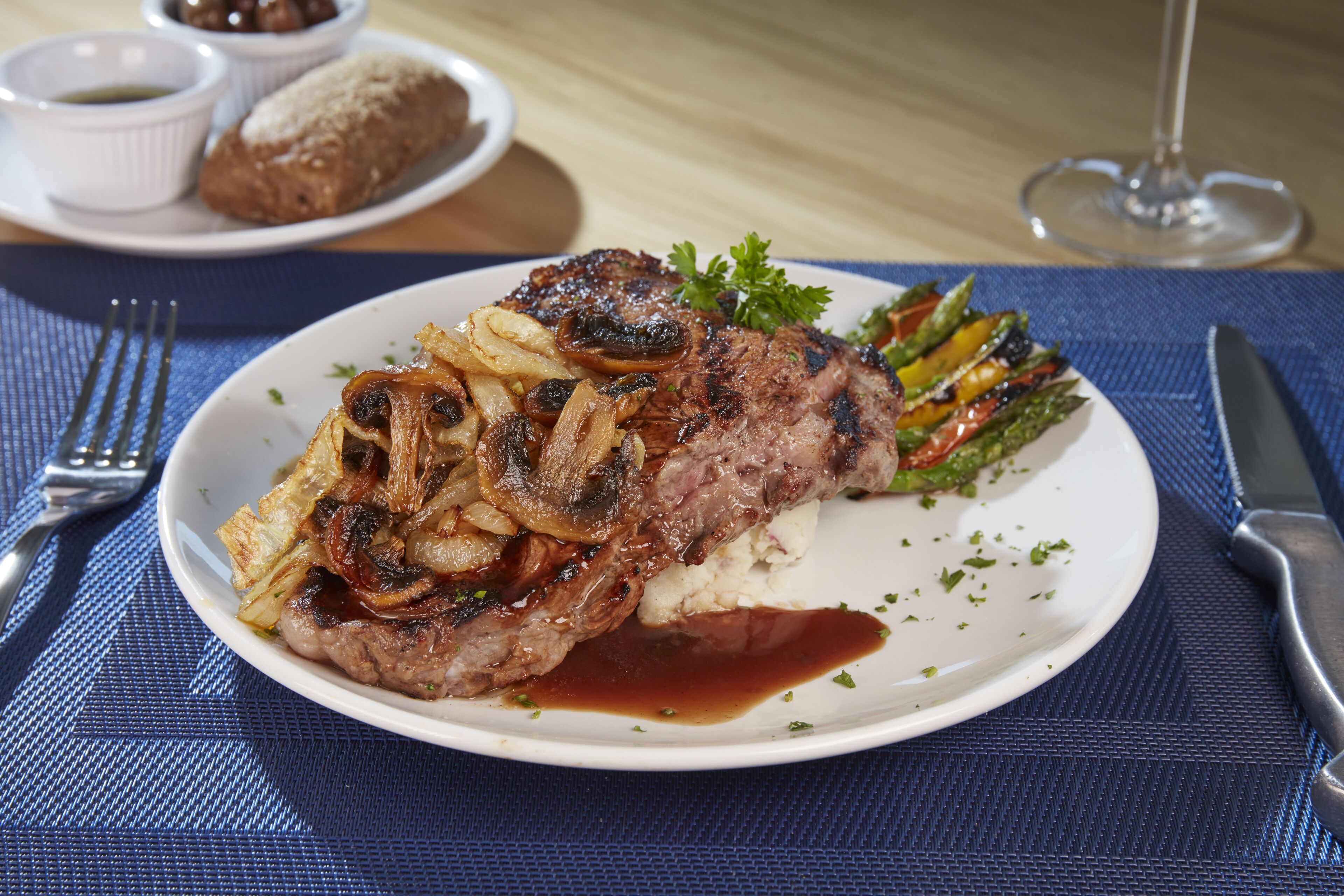 A plate of steak with mushrooms and a side of vegetables, accompanied by bread and a glass of wine on a table.