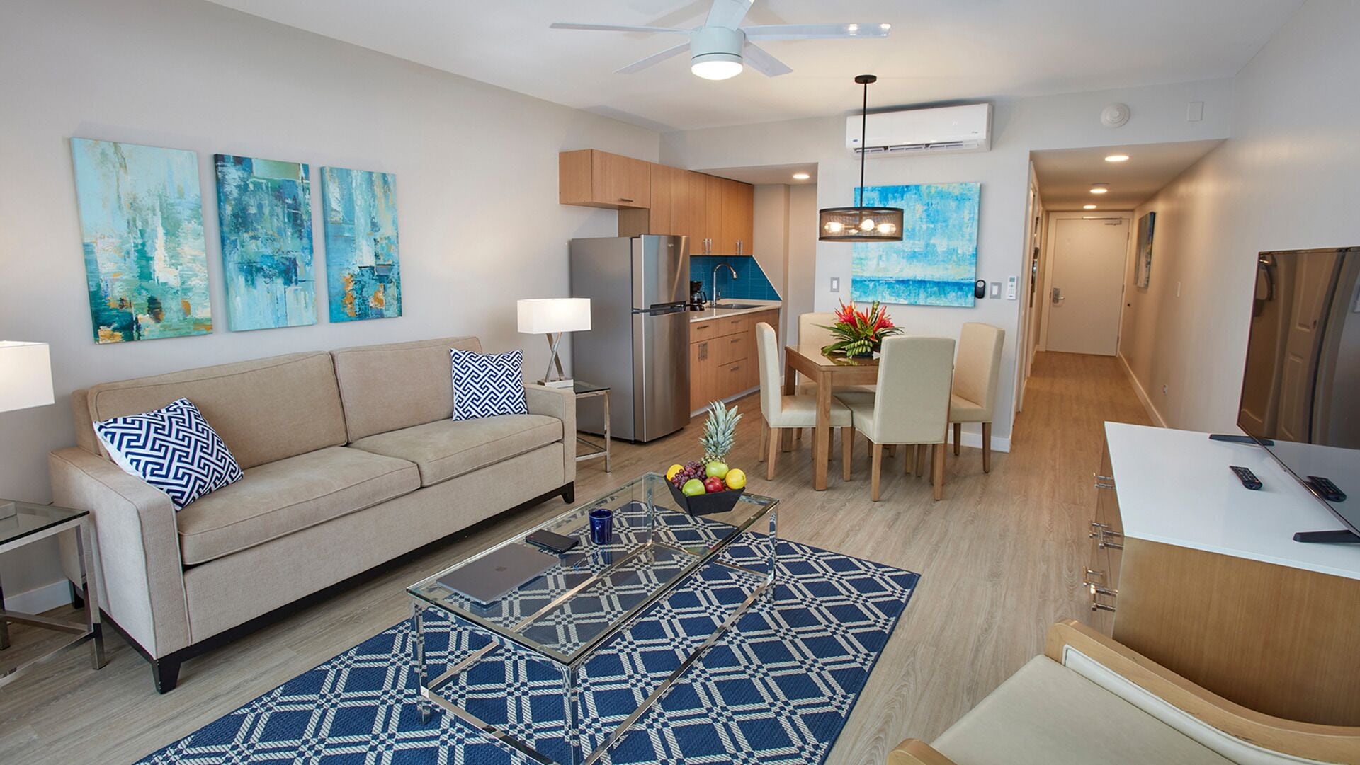 A modern living room with a sofa, coffee table, and artwork. A kitchen with a refrigerator, stove, and dining area is visible in the background.