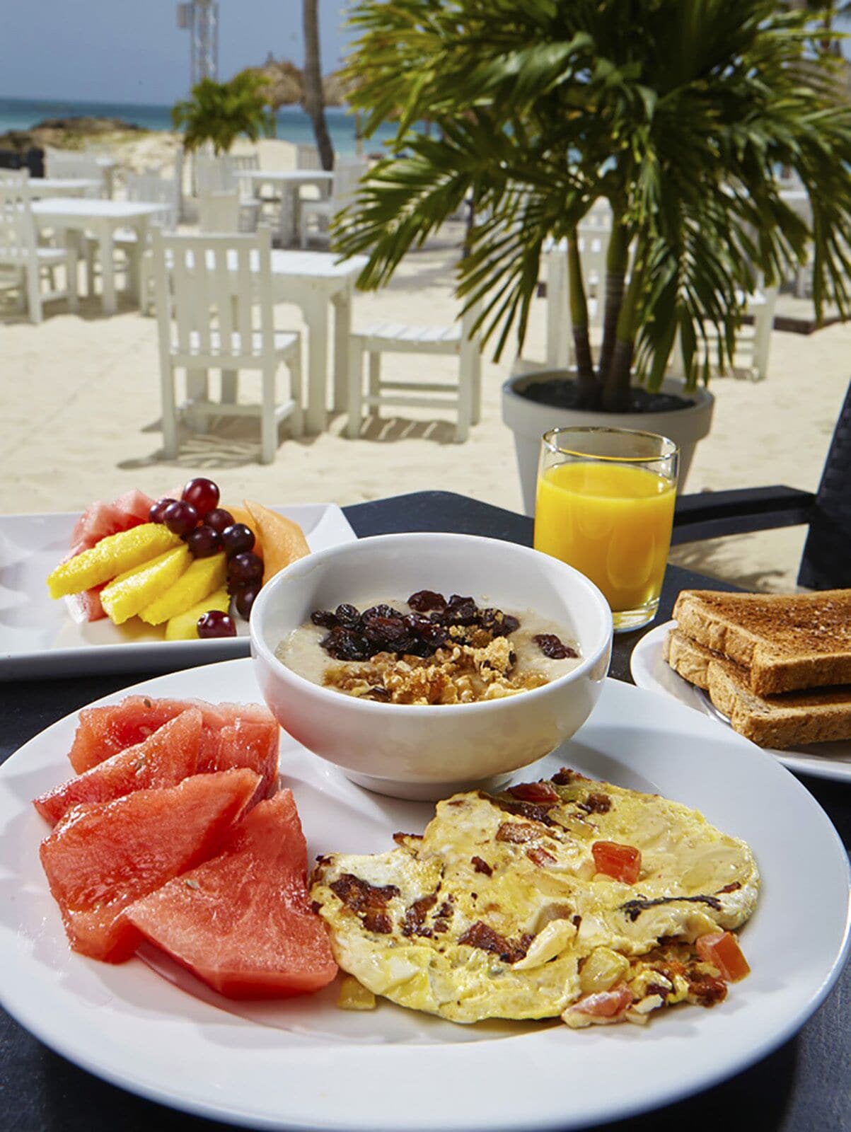 A breakfast setting with a bowl of cereal, toast, omelette, watermelon, and a glass of orange juice on a table at a beachside location.