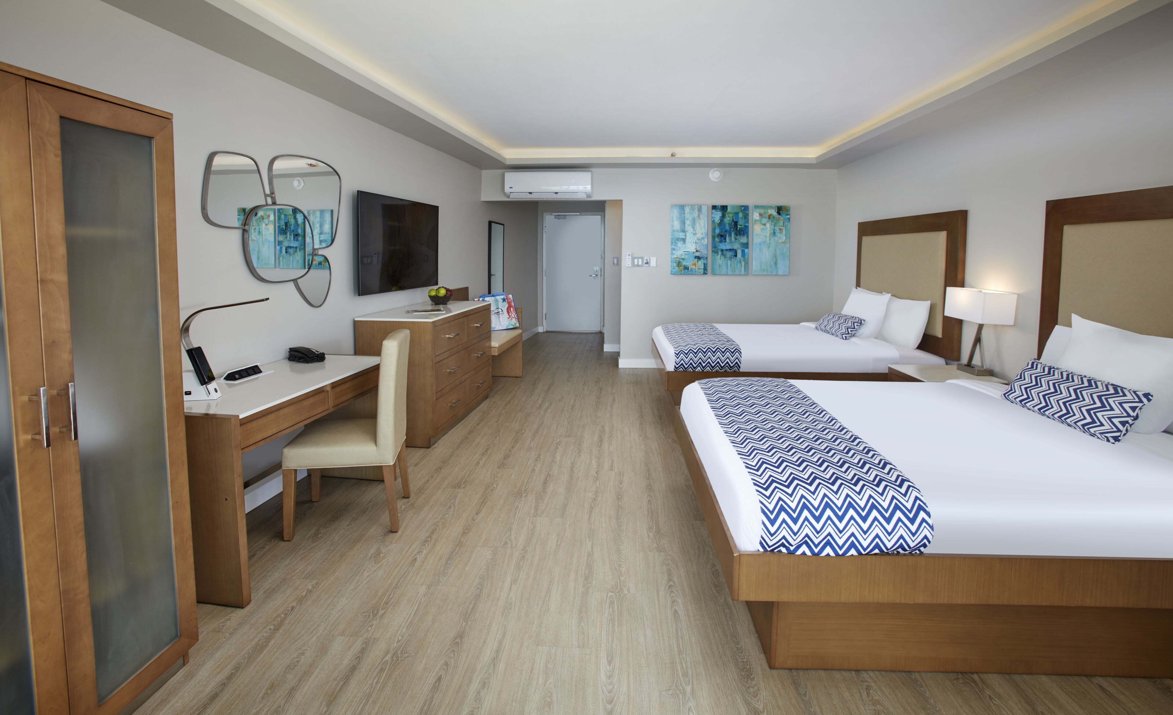 A hotel room with two beds, a desk, a chair, a TV, and artwork on the walls.