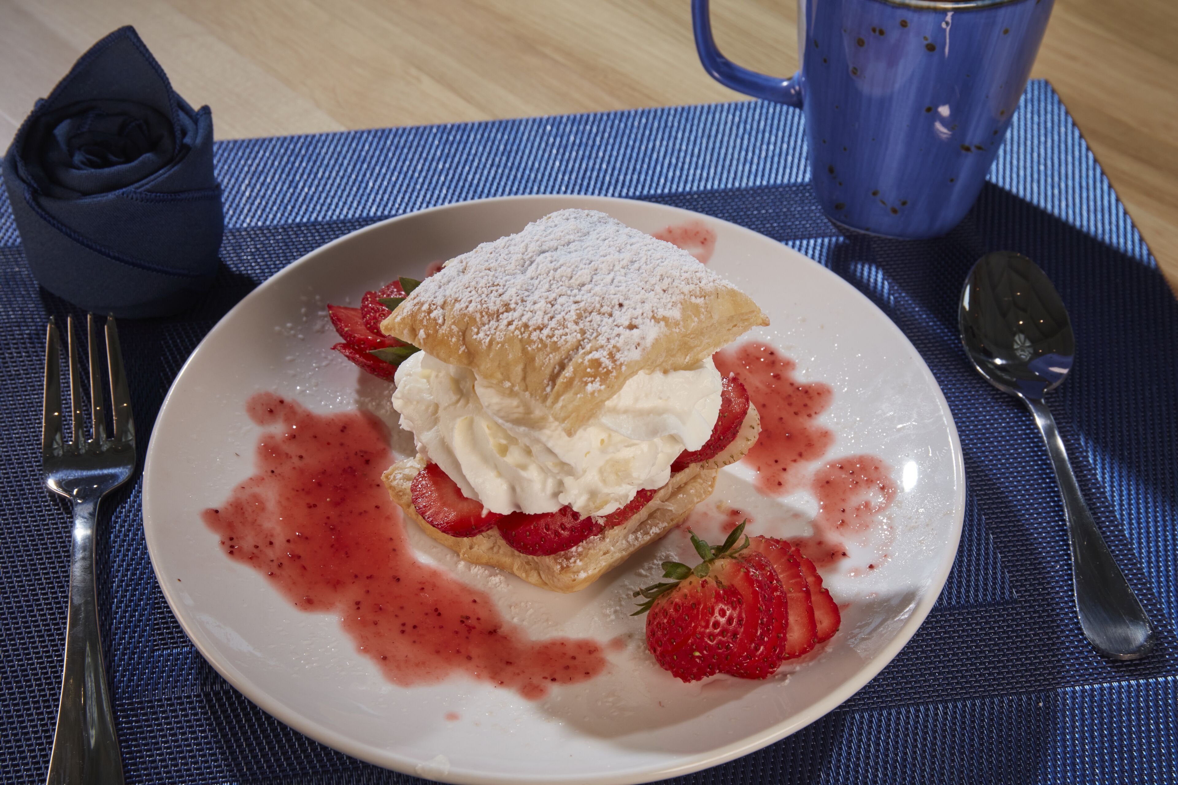Une assiette à dessert avec une pâtisserie, de la crème fouettée et des fraises, le tout posé sur un set de table bleu.