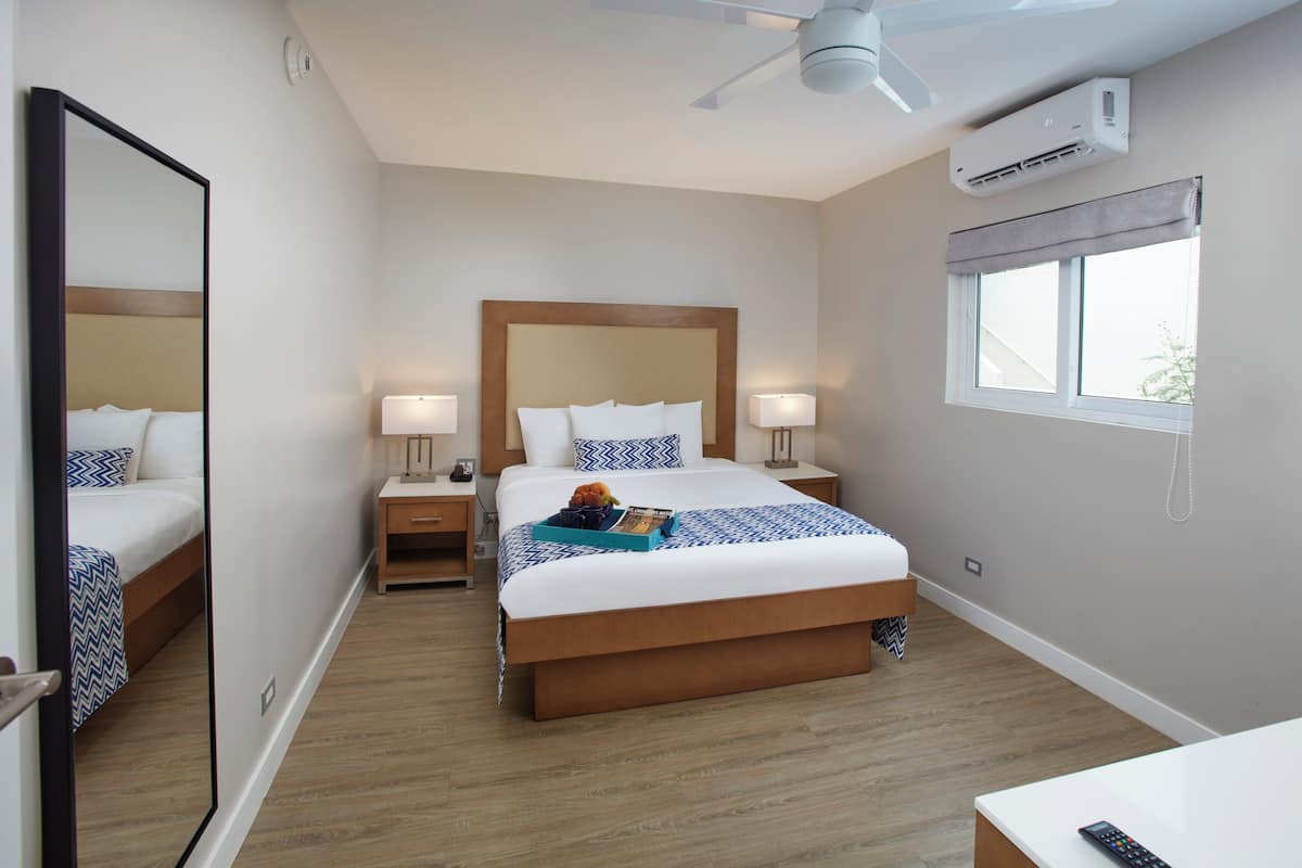Une chambre d’hôtel comprenant un lit, des tables de chevet, un ventilateur de plafond, un appareil de climatisation, une fenêtre avec des stores et un miroir.