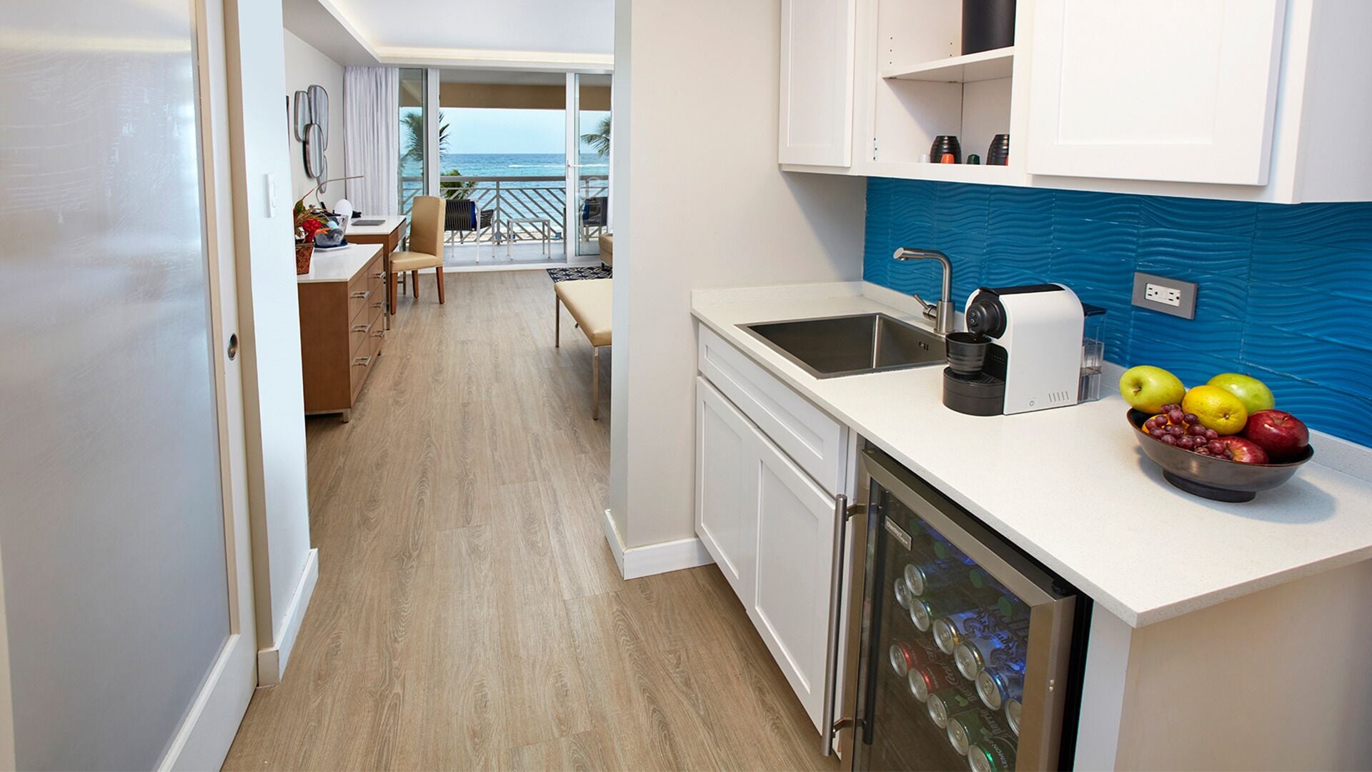 A modern kitchen with a view of the ocean, a dining area with a bench, and a refrigerator with a fruit bowl.