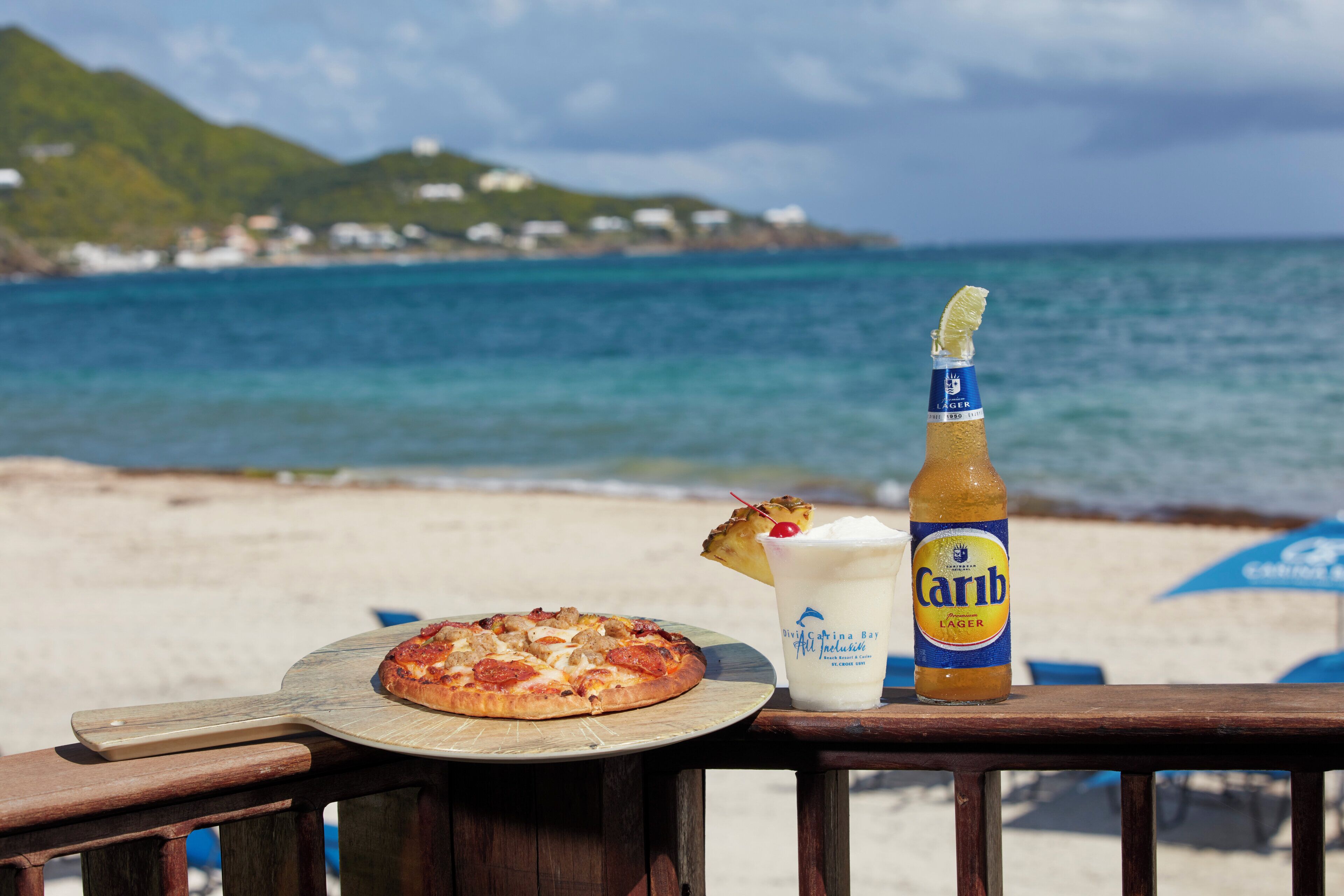 Une table installée sur la plage, avec une pizza, une boisson et une bouteille de bière.