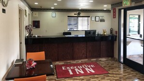 Zona de recepción con un mostrador de madera, una computadora y un tapete rojo con el texto "Executive Inn". Se observa una ventana con persianas, un reloj y un cartel en la pared.