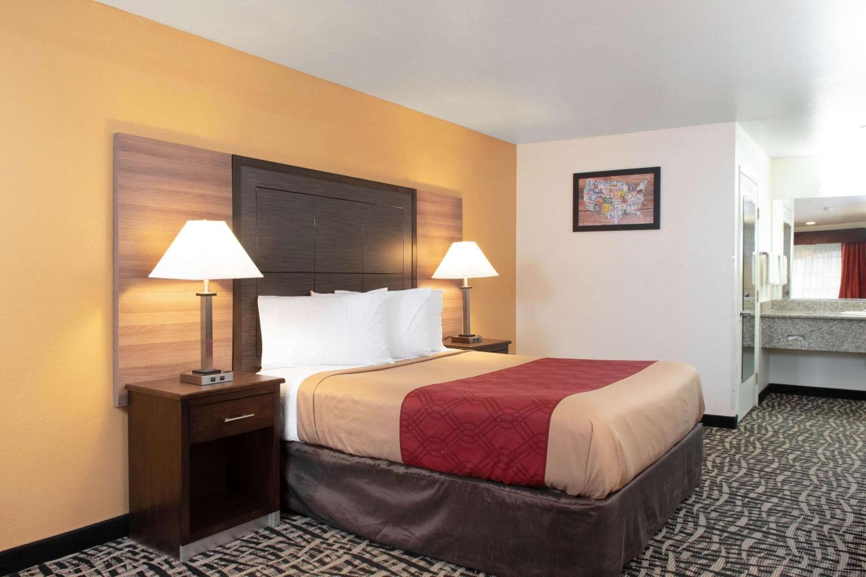 A hotel room with a large bed, two bedside tables with lamps, a wooden headboard, a small desk with a chair, a bathroom mirror, and a patterned carpet.