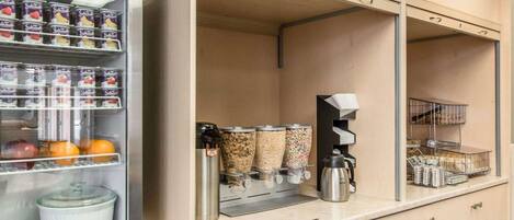 Un área de desayuno autoservicio con un refrigerador, máquina de café y diversos recipientes de comida.