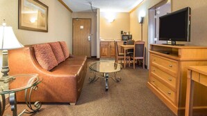 A compact hotel room with a brown leather sofa, a glass coffee table, a TV on a wooden stand, a small kitchenette, and a door leading to another room. - Quality Inn & Suites Casper near Event Center (Casper)