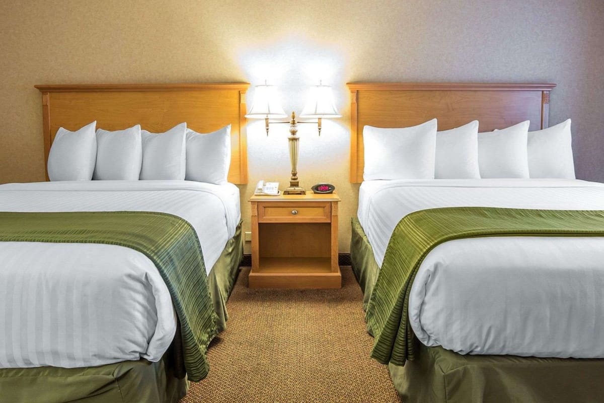 A hotel room with two beds, each with white linens and green bedspreads. There is a wooden headboard, a nightstand with a lamp, and a telephone.