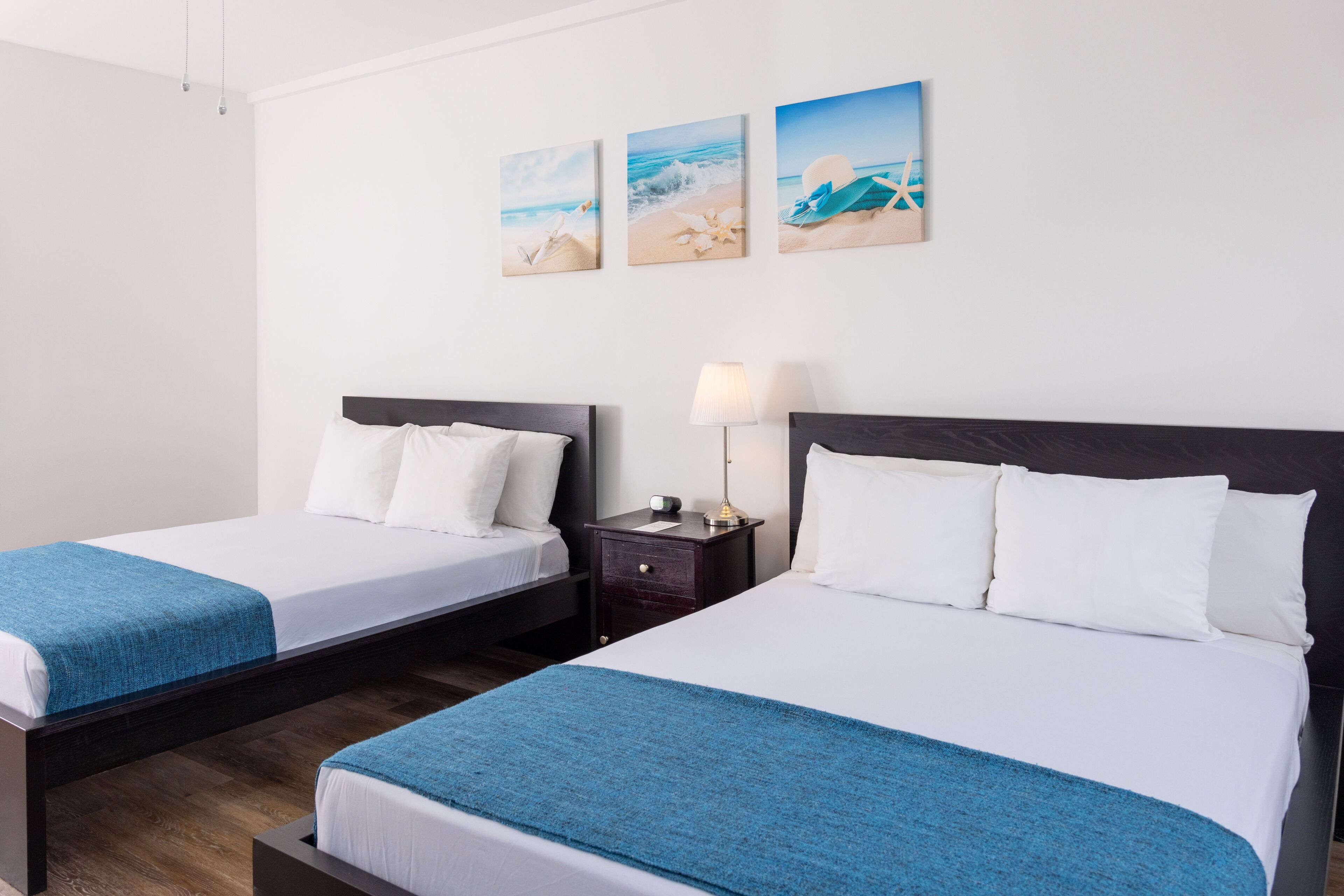 A hotel room with two single beds, a nightstand with a lamp, and three beach-themed wall art pieces.