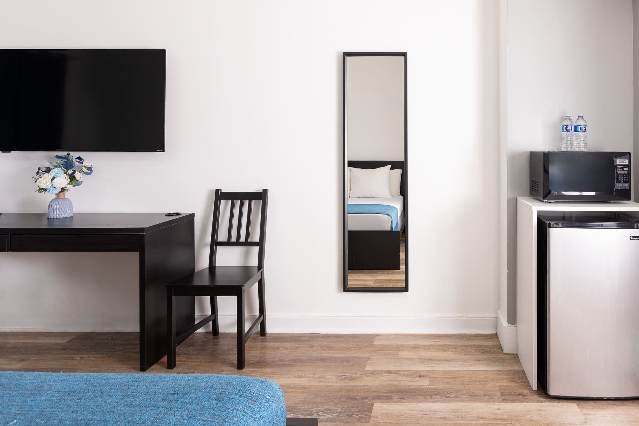 A hotel room with a flat-screen TV, a desk with a vase of flowers, a chair, a bed with a white pillow, a mirror, and a minibar.
