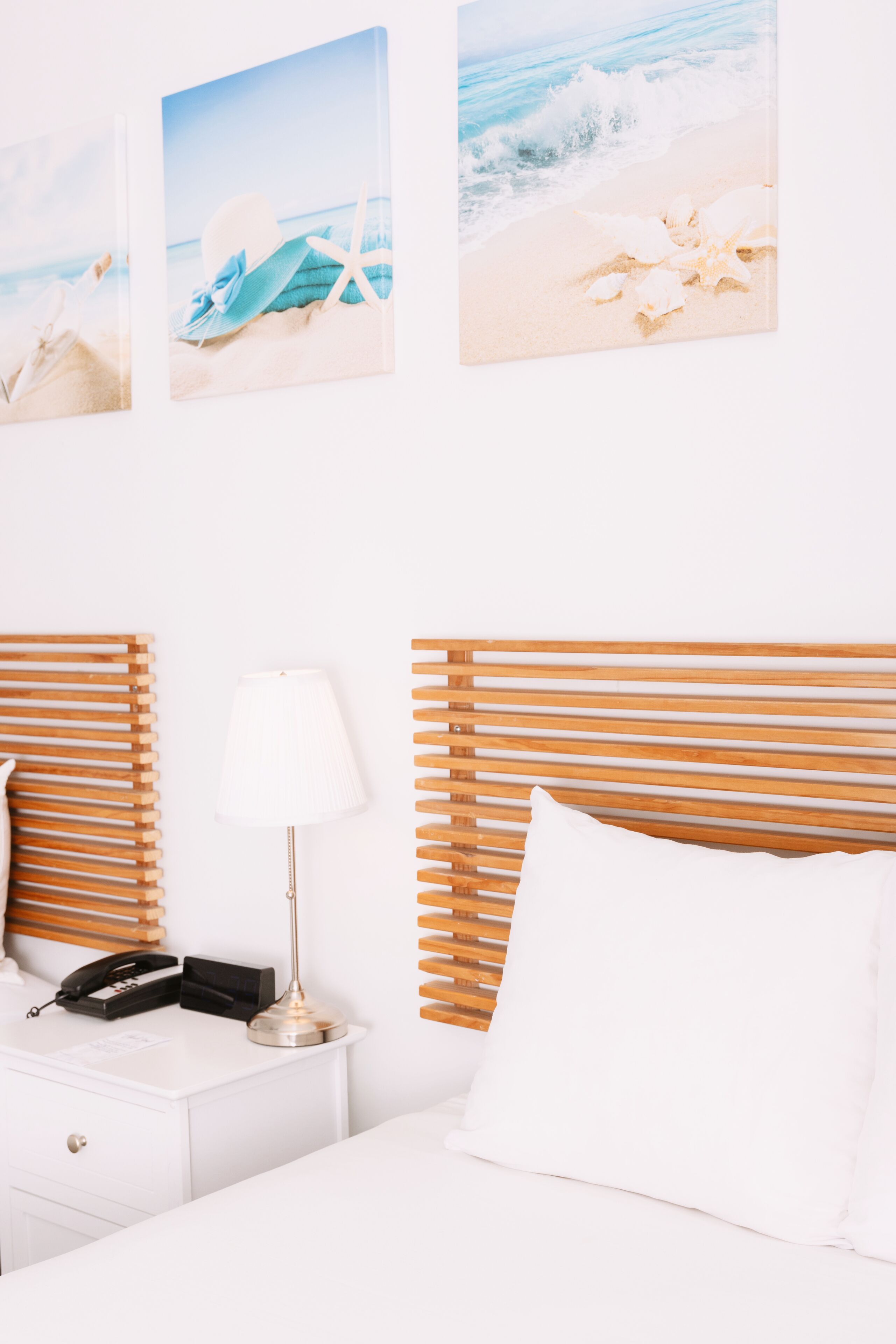 Habitación de hotel con cabecera de madera, ropa de cama blanca, lámpara de noche, teléfono y tres obras de arte con motivos playeros.