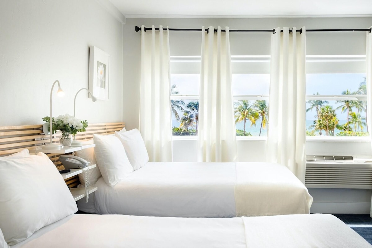 Une chambre d’hôtel avec deux lits, une table de chevet avec une lampe, un tableau au mur et une vue sur les palmiers et l’océan, visible à travers de grandes fenêtres.