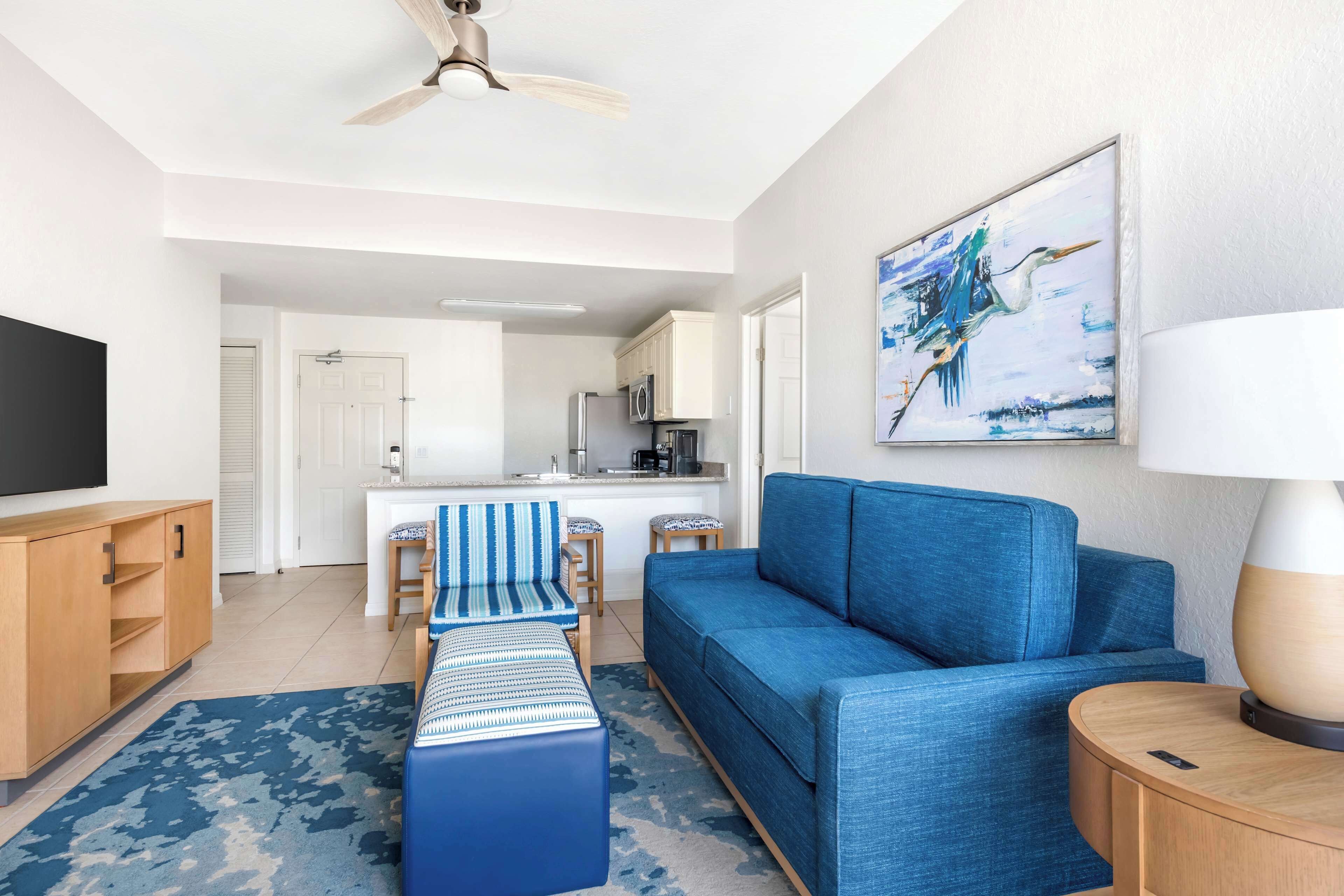 A modern living room with a blue sofa, a coffee table, and a TV on a wooden cabinet. There is a kitchen in the background with a microwave and a refrigerator.