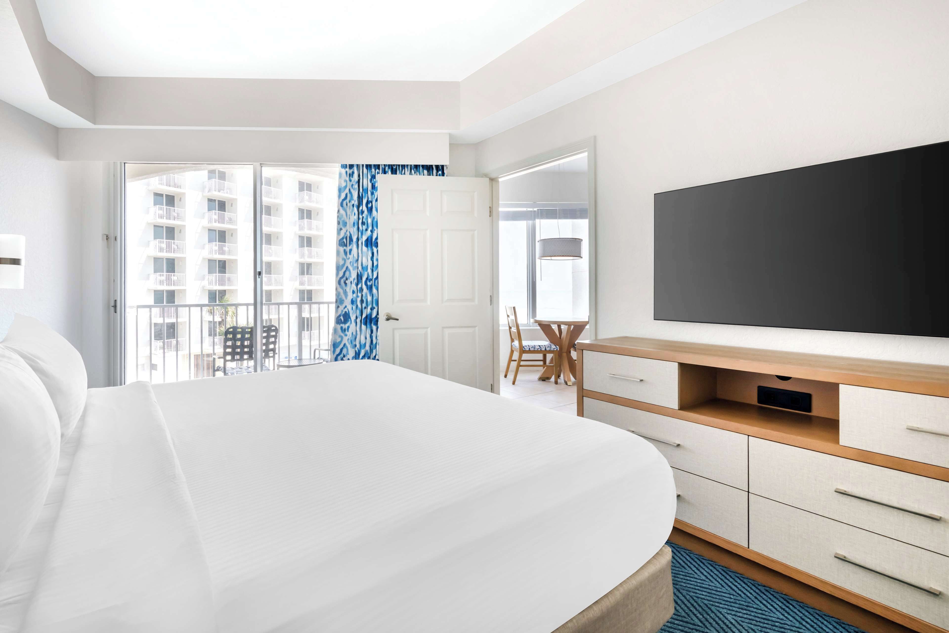 A hotel room with a large bed, a TV, a desk, and a balcony with a view of buildings.