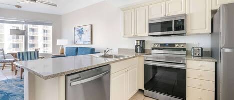 A modern kitchen with a dishwasher, microwave, oven, and refrigerator. There is a dining area with a blue sofa and a coffee maker.