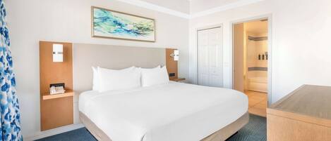 Une chambre d’hôtel avec un grand lit, un ventilateur de plafond, un tableau, une table de chevet et une salle de bain visible à travers une porte ouverte.