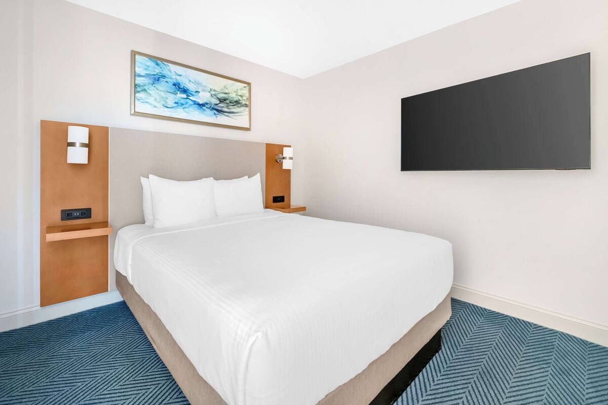 Ein Hotelzimmer mit einem großen Bett, einem Gemälde an der Wand und einem Flachbildfernseher.